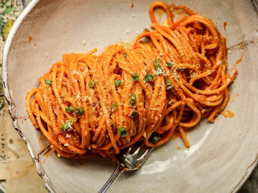 A bowl of vegan gochujang vodka pasta tossed in a rich red tomato sauce, garnished with green herbs and grated cheese. A fork rests beside the pasta, ready to twirl the strands. The dish is served in a rustic, shallow ceramic bowl.